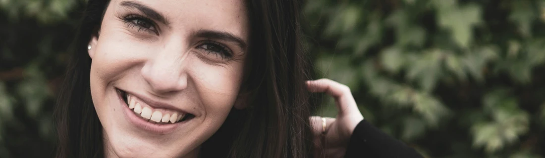 close up on a beautiful woman with long brown hair is smiling wide in front of a bush